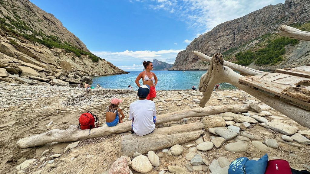 Am steinigen Ufer einer abgelegenen Bucht, während ein Jens und die Kinder auf einem Baumstamm sitzen. Im Hintergrund türkisblaues Meer, Felsen und andere Badegäste.