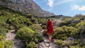 Silke mit dem großen roten Rucksack wandert auf einem felsigen Pfad bergauf, umgeben von dichtem Grün und Bergen