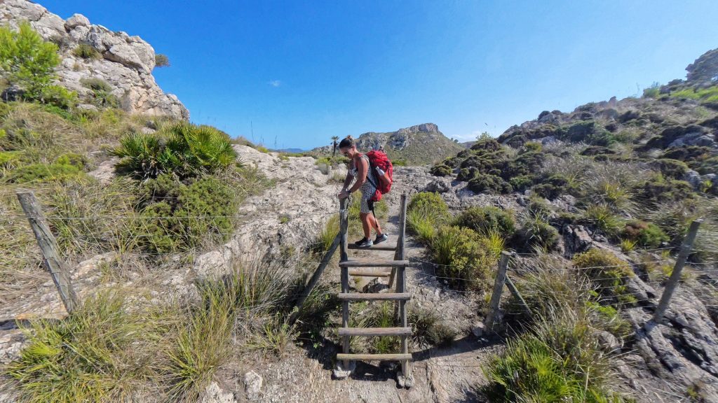 Frau klettert über ein Gatter auf einer Wanderung im Nationalpark Llevant auf Mallorca, begleitet von Familie