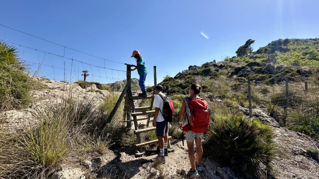 Kind klettert über ein Gatter auf einer Wanderung im Nationalpark Llevant auf Mallorca, begleitet von Familie