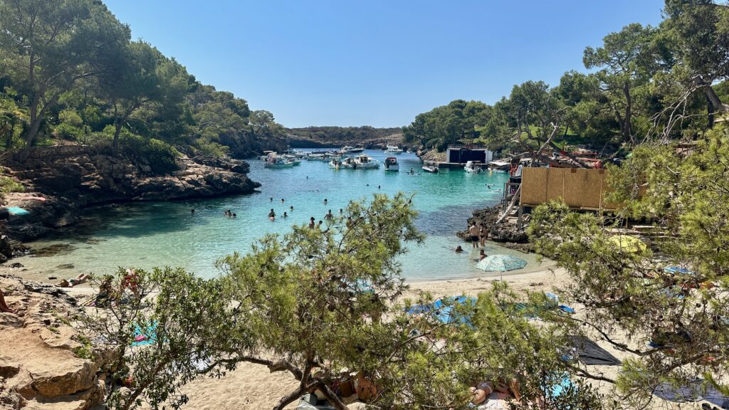 Zwischen Bäumen versteckt die Mini Bucht mi türkisblauen Wasser und Segelbooten im Hintergrund