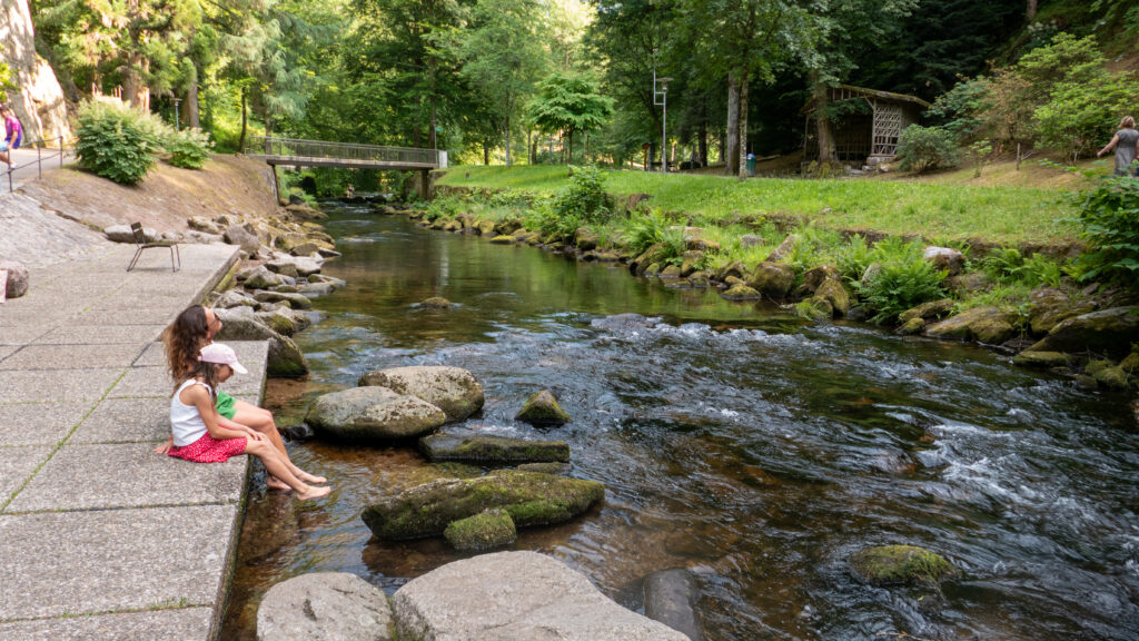 Mutter und Kind sitzen an der Enz im Kurpark Bad Wildbad mit den Füßen im Wasser, Steine und Farne am Ufer