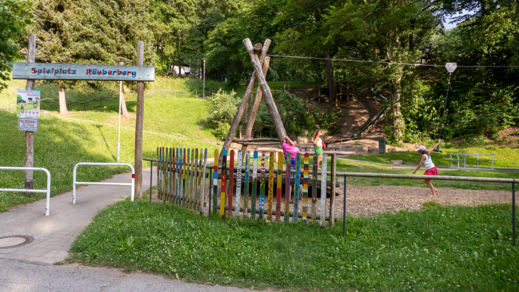 Eingangsschild Spielplatz Räuberberg im Kurpark Bad Wildbad mit buntem Holzzaun und Seilrutsche im Hintergrund